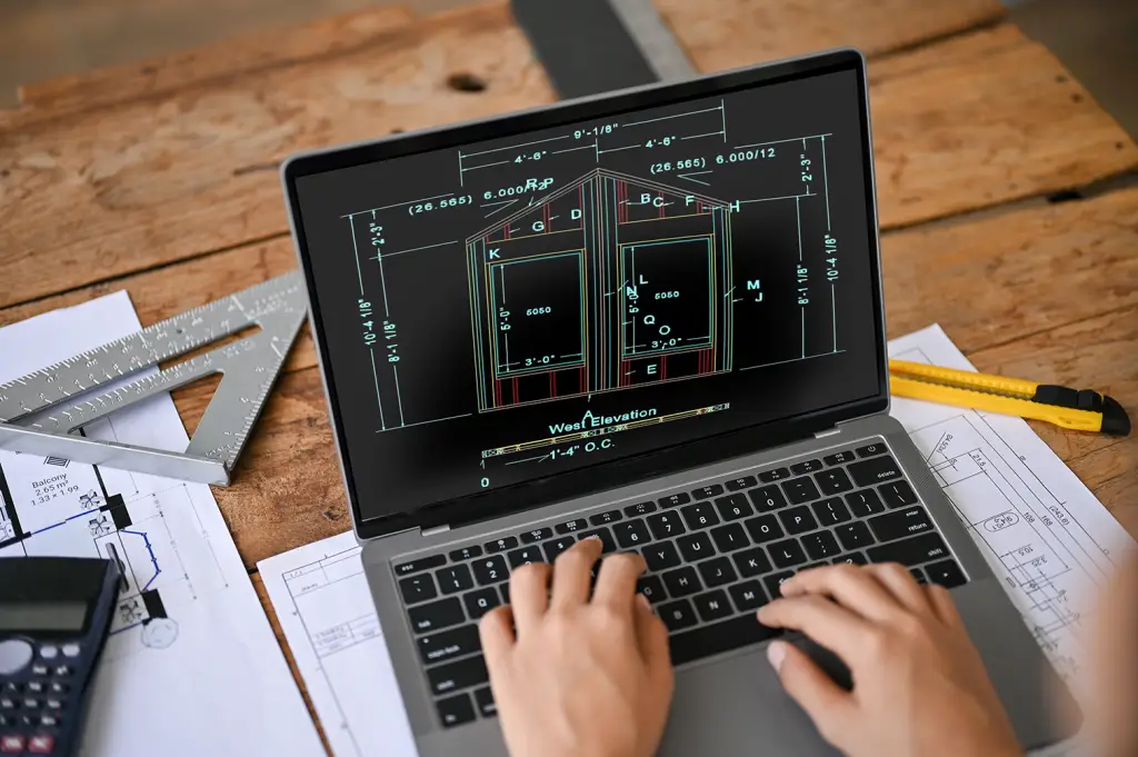 A laptop on a wooden desk displays a detailed architectural “West Elevation” drawing with measurements and labeled sections. Around the laptop are drafting tools including a metal triangle ruler, calculator, printed blueprints, and a yellow utility knife.